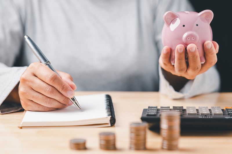 © Mdisk - stock.adobe.com Foto: Hand mit Notizbuch und Sparschwein